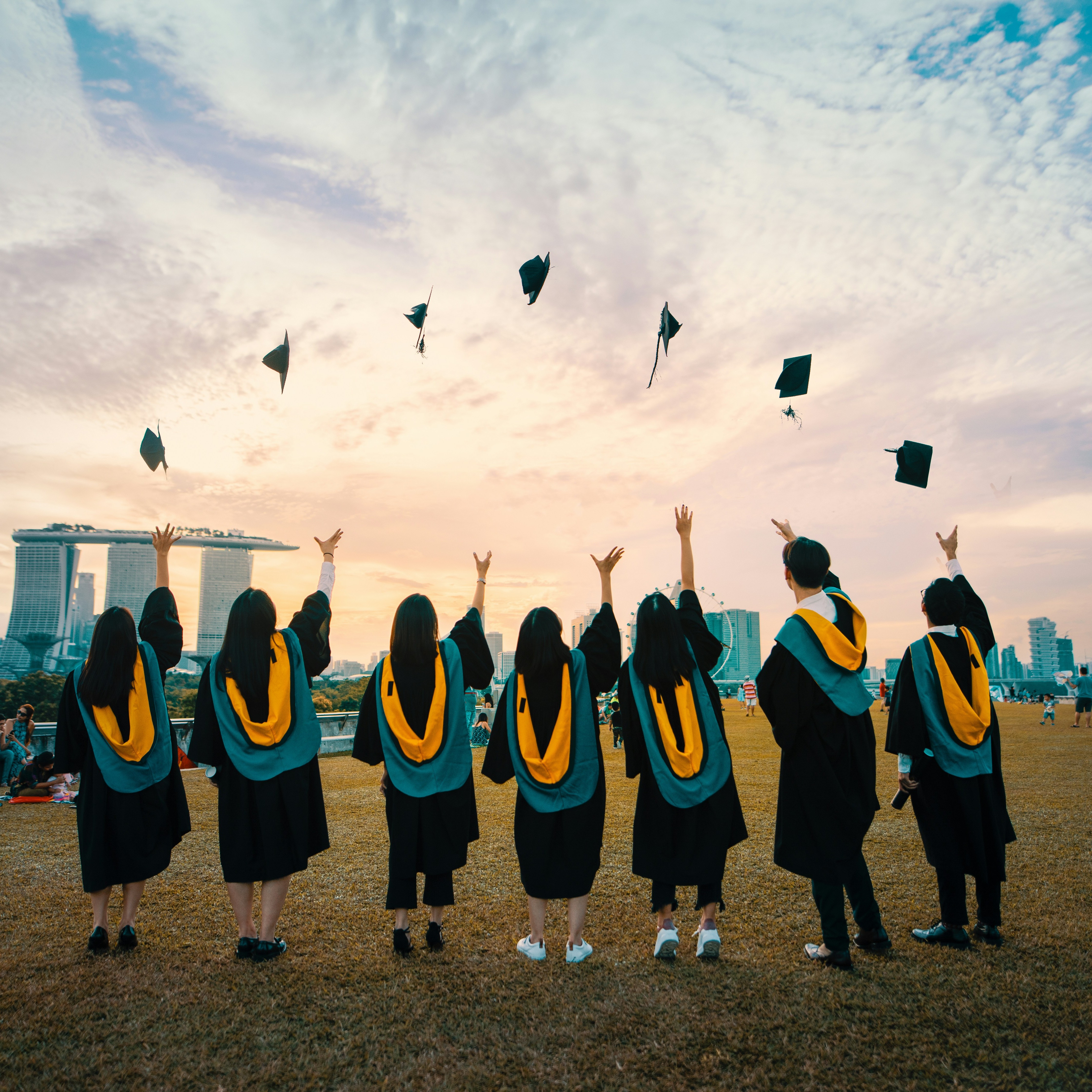 A group of seven students in their graduation attire tossing their caps in celebration while in a field looking towards the open sky.