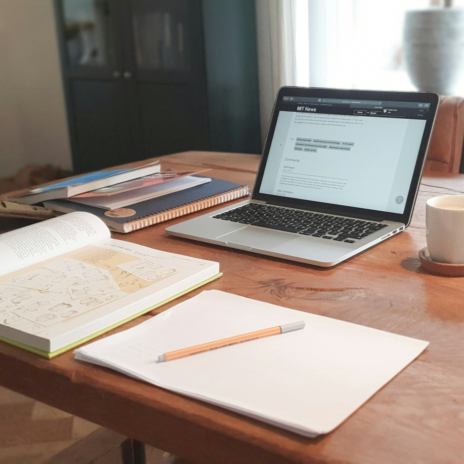 A laptop, textbook, a few notebooks, pen and a coffee mug resting on a desk. A set up for a nice study session.
